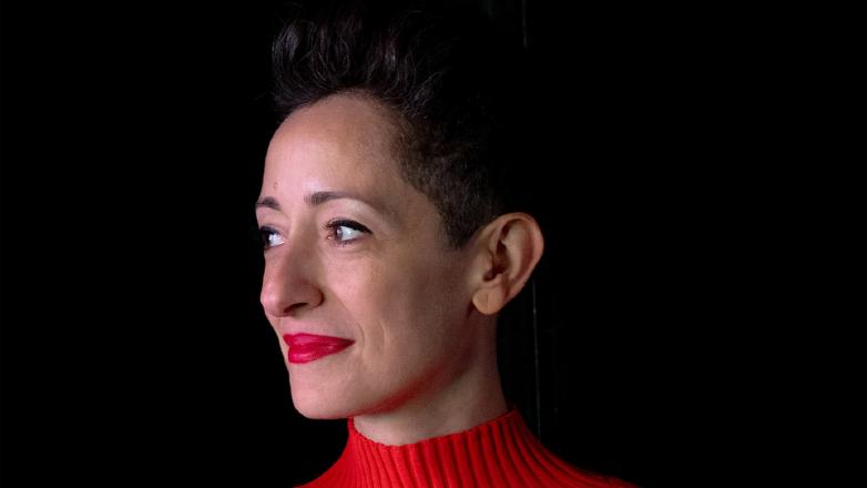 A woman with dark hair wears a red turtleneck and red lipstick against a black background.