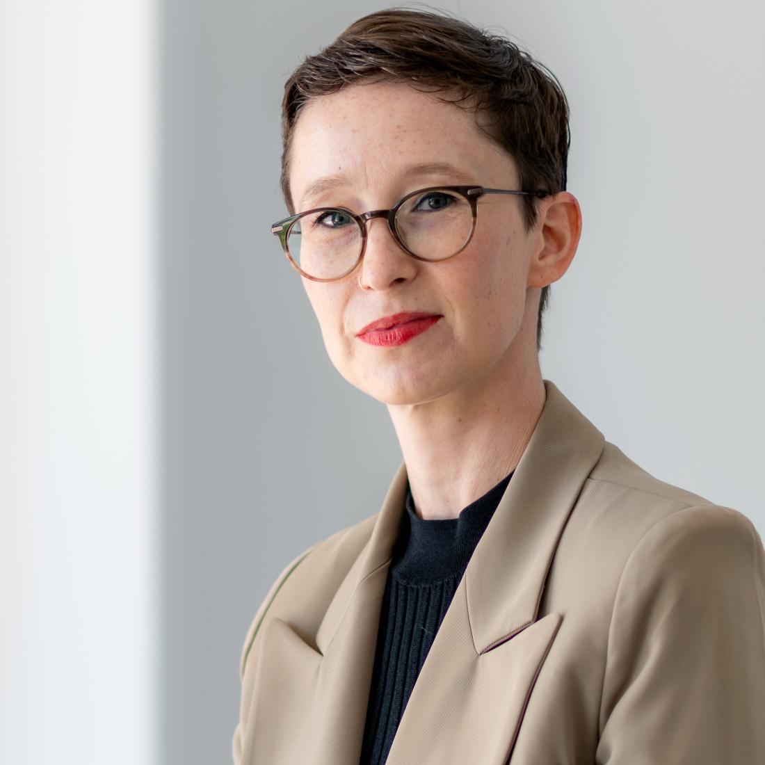 headshot photo of woman wearing khaki jacket, glasses, with short dark hair