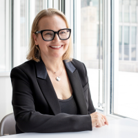 headshot of blond woman sitting at a desk, wearing black suit and black frame glasses