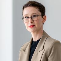 headshot photo of woman wearing khaki jacket, glasses, with short dark hair