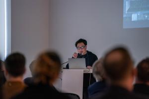 Image of man standing at a lecturn projecting images on a wall and speaking to a crowd